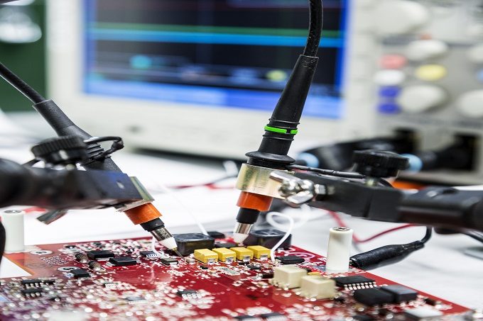 Close-up of two probes held on an electronic circuit card with an oscilloscope in the background.